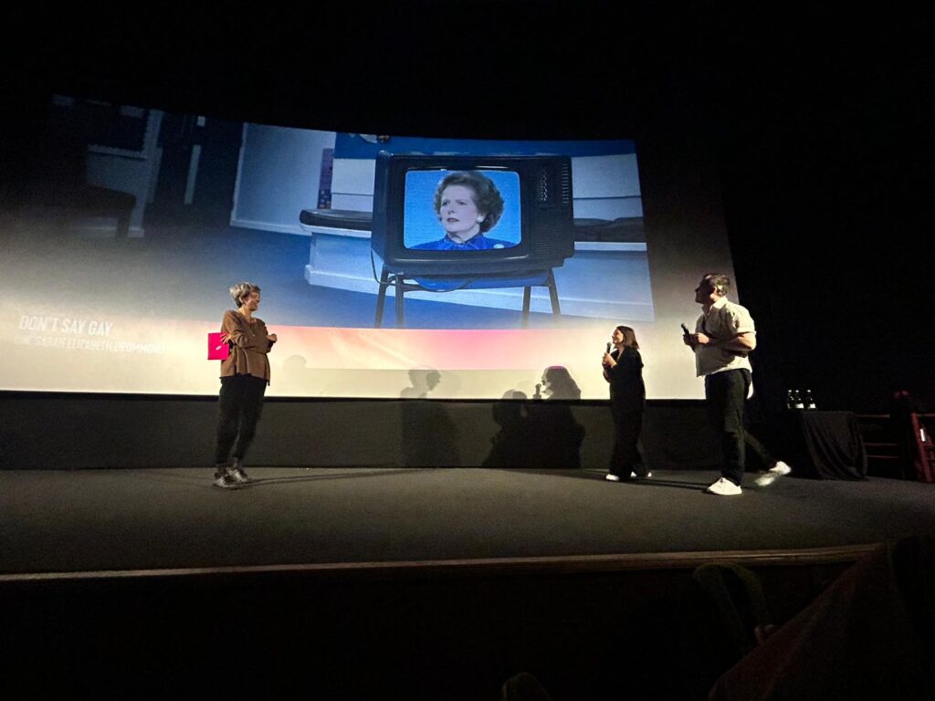 Three people in front of a screen with a tv on margaret thatcher being projected